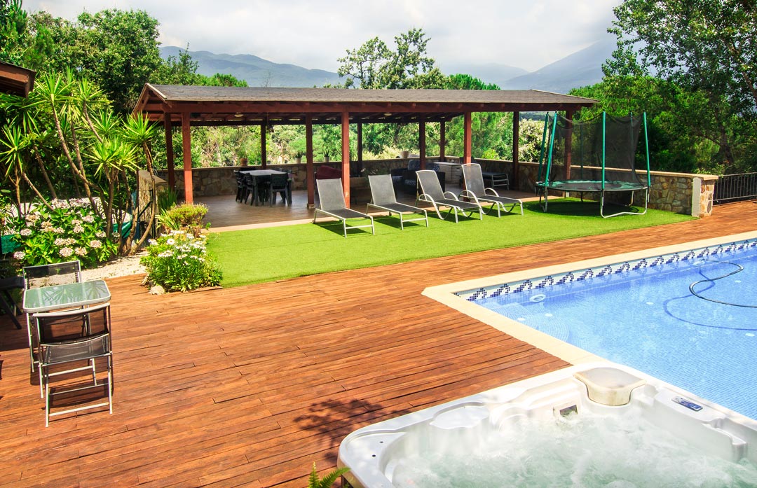 A spacious outdoor area features a salt-water pool alongside a wooden deck. Sun loungers are arranged for relaxation, while a covered porch provides shaded seating. A trampoline is visible on the green lawn, surrounded by lush greenery and mountain views in the background.