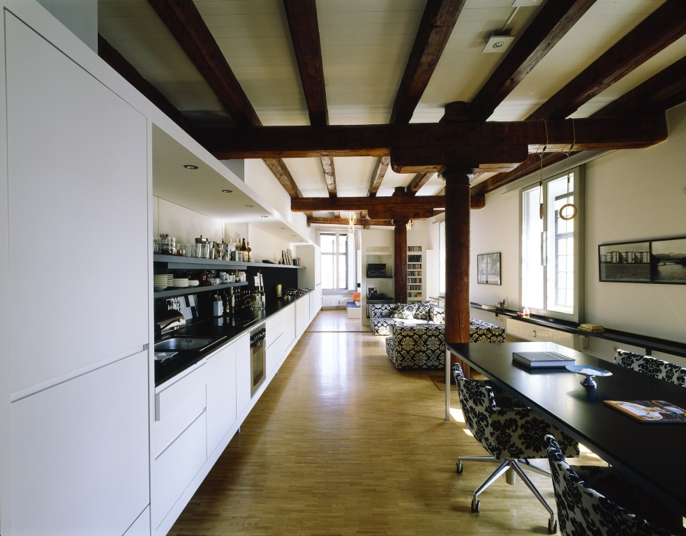 The spacious loft features high wooden beams and an open layout. A sleek kitchen runs along one side, equipped with modern appliances. A large dining table sits alongside patterned chairs, while a cozy seating area is arranged for relaxation. Ample natural light enters through large windows.