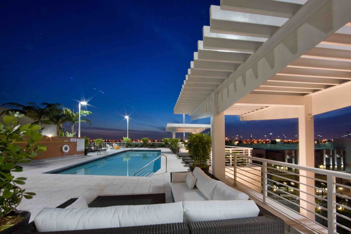 The rooftop area features a pristine swimming pool illuminated by soft lighting. Comfortable lounge seating is available under a shaded pergola, providing a relaxing spot to enjoy the evening views. Palm trees are visible in the background, enhancing the tranquil atmosphere.