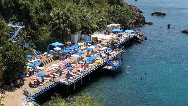 A sunlit beach area features colorful umbrellas and lounge chairs arranged on a wooden deck by the water's edge. People can be seen swimming and relaxing, while rocky cliffs serve as a backdrop, enhancing the coastal scenery.
