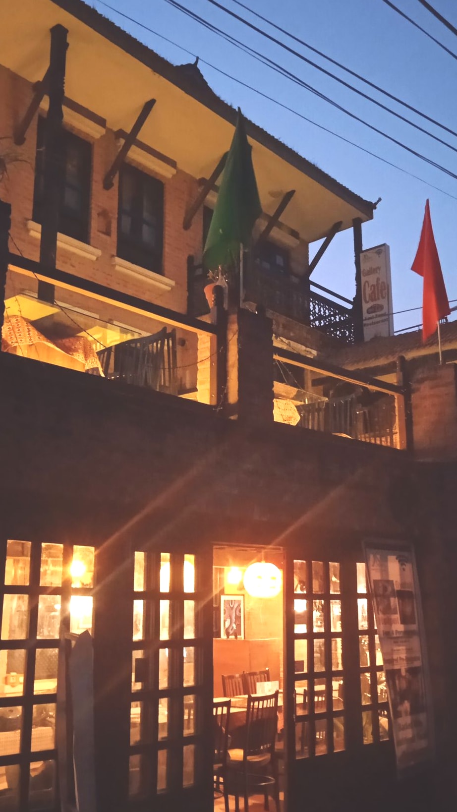 The exterior of a multi-story building is illuminated by warm lights, creating a welcoming ambiance. Balconies adorned with potted plants and flags are visible above a cozy café entrance, which features large glass doors and a rustic wooden design, inviting guests to explore.