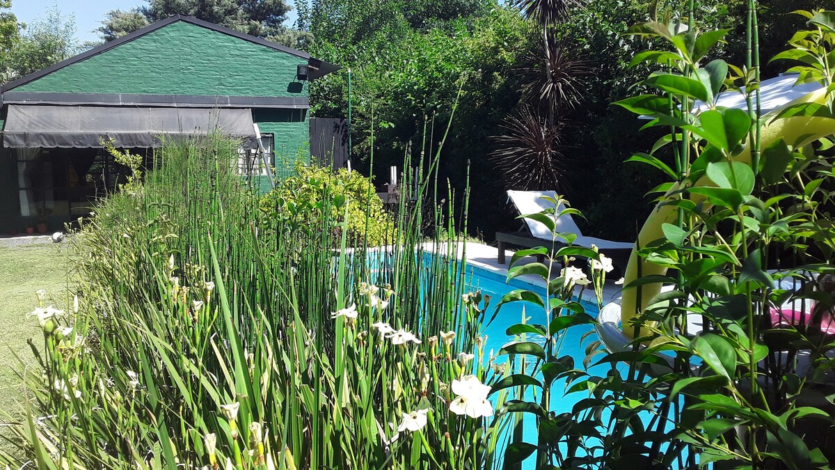 A well-manicured garden features lush greenery and flowering plants alongside a clear swimming pool. A shaded area and lounge chairs invite relaxation. The background reveals a charming green building nestled among trees, enhancing the tranquil outdoor setting.