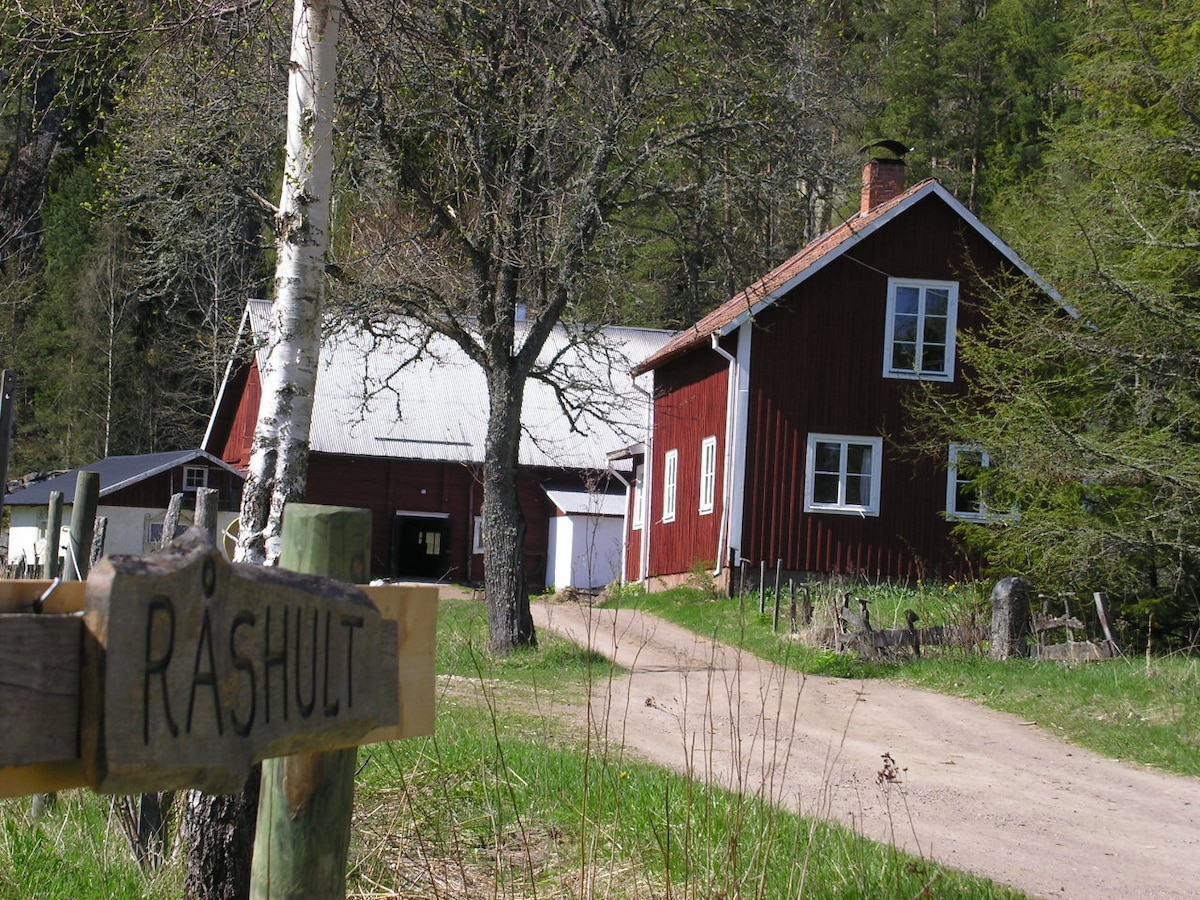 A charming red house, featuring white trim, is positioned near a gravel path lined with green grass. Trees surround the area, providing a natural setting. A sign marked 'Råshult' is visible in the foreground, indicating the property name.