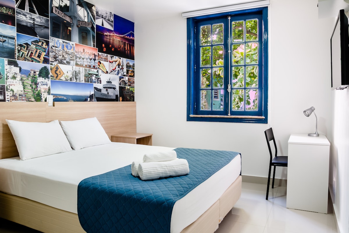A well-appointed room features a spacious queen-size bed with a blue quilt and neatly folded towels. A wooden desk with a lamp and a black chair are present. The wall is adorned with a vibrant collage of images, and large windows with blue frames allow natural light to enter.