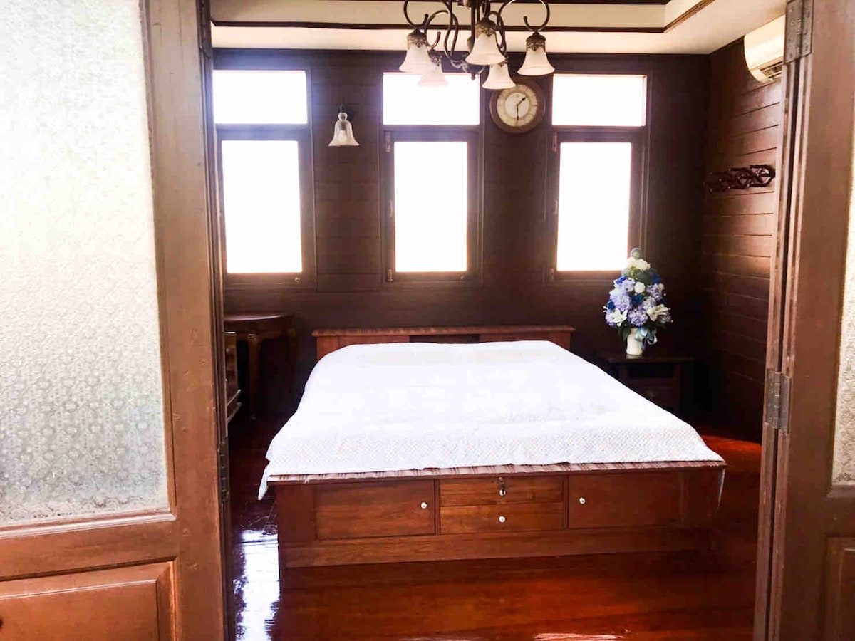 A spacious sleeping area features a wooden bed with a decorative blue and white quilt. Natural light enters through multiple windows adorned with curtains, illuminating the room. A clock and a floral arrangement are positioned on the walls, enhancing the room's peaceful ambiance.