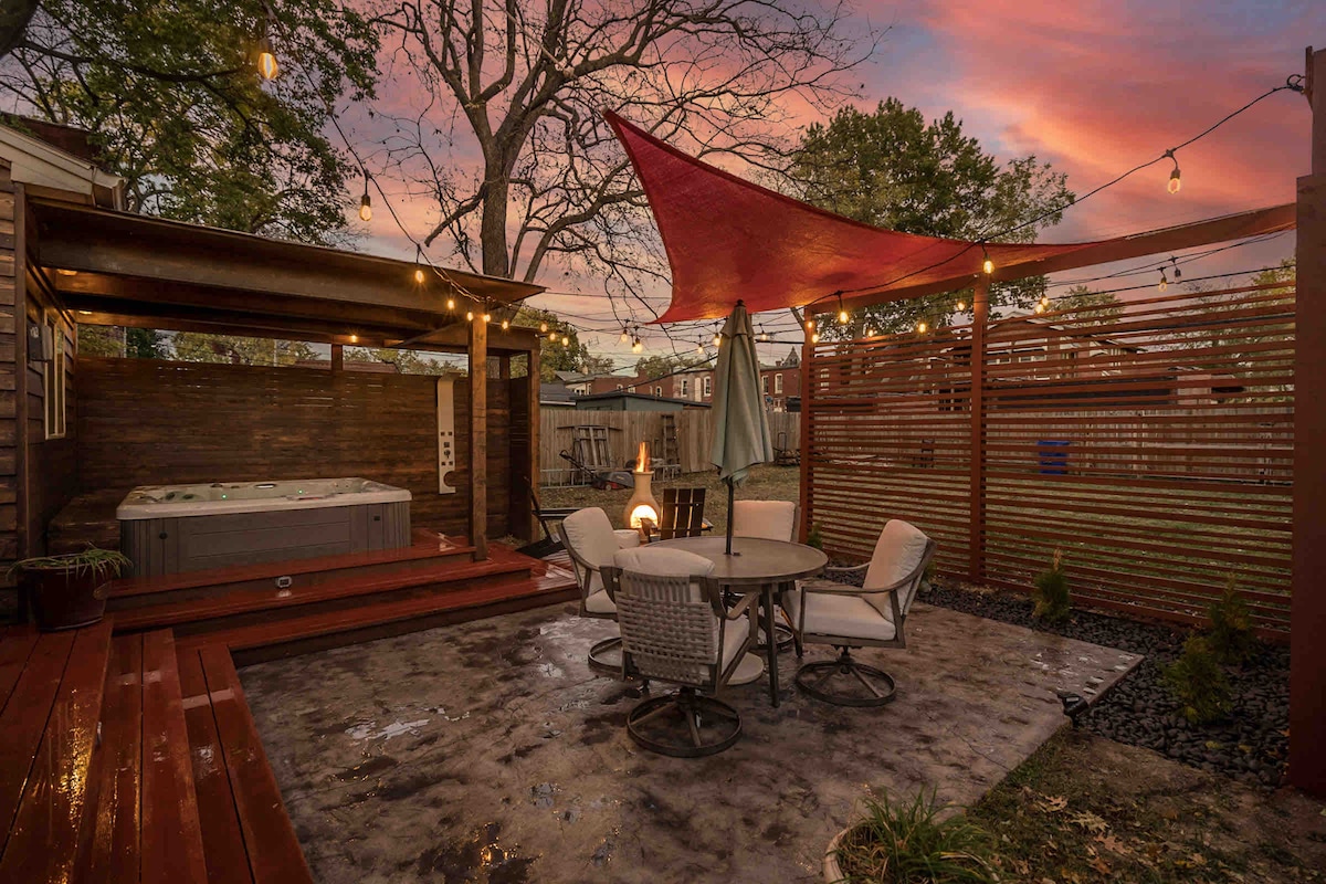 An outdoor patio space is depicted featuring a hot tub beneath a wooden canopy. A set of outdoor dining furniture, including a round table and four chairs, is positioned nearby. String lights are hung above, adding ambient lighting, while a red canopy provides shade.