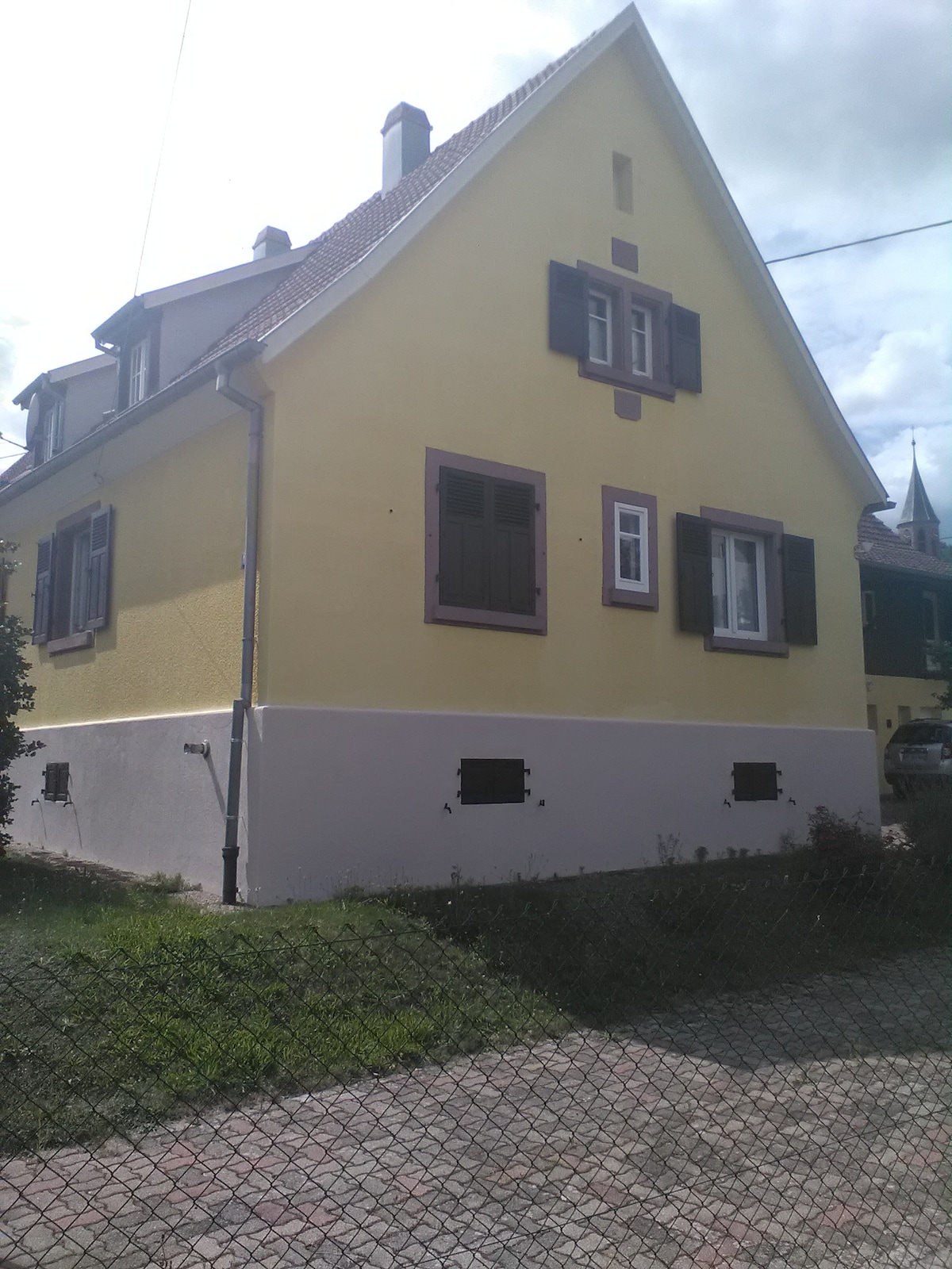 The charming yellow house features a distinctive architectural style with dark shutters framing the windows. The exterior is divided by a lighter-colored base, and the lawn is well-kept, providing a welcoming view of the property.