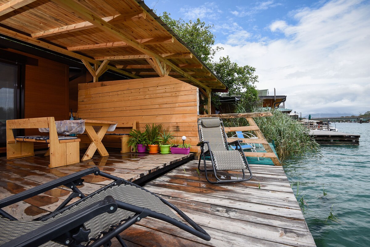 A wooden terrace extends over the water, featuring a wooden table and chairs. A pair of lounge chairs are positioned for relaxation. Lush greenery is visible nearby, alongside a wooden structure that provides shelter.