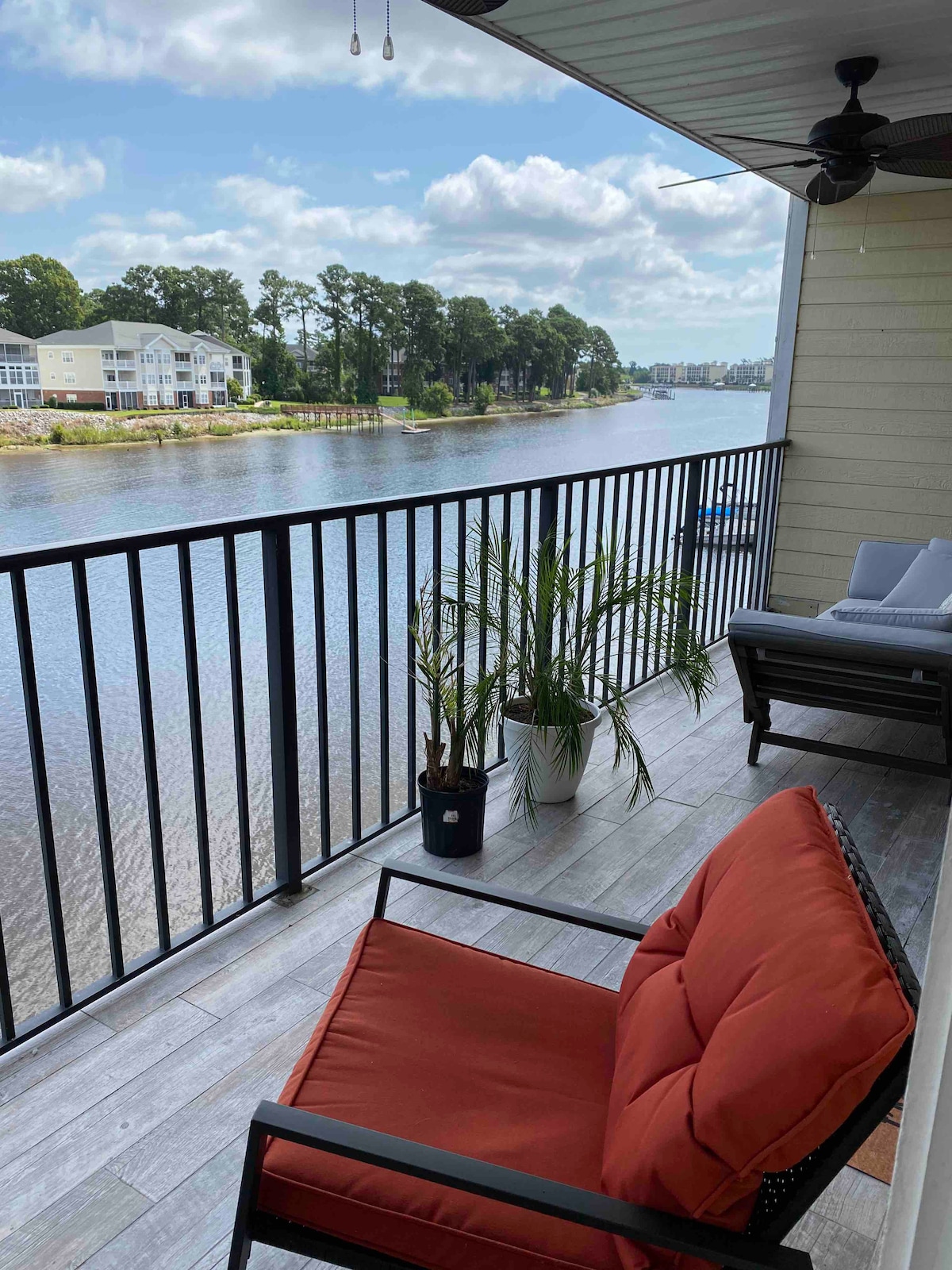 A spacious balcony overlooks the river, featuring a comfortable orange chair and a potted plant. The railing provides an unobstructed view of the tranquil water, while trees line the opposite bank, creating a serene environment for relaxation.