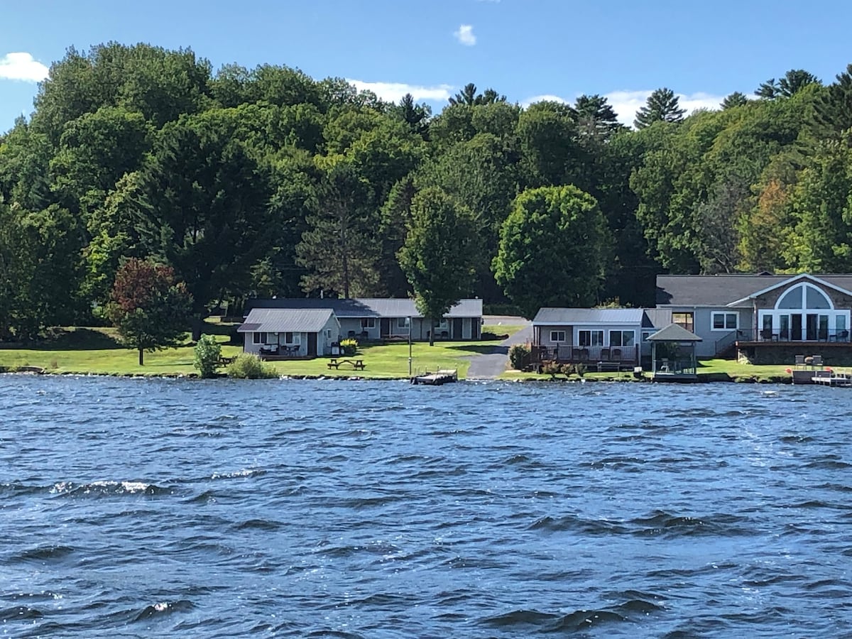 A line of cabins is positioned along the shore of Portage Lake, surrounded by greenery and trees. Picnic tables are visible on the manicured lawn, and the calm water reflects the blue sky. The scene presents a peaceful lakeside setting for relaxation.