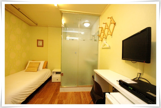 A single bed is positioned against a light green accent wall, featuring a minimalist design. A glass-enclosed shower area is visible, offering a modern touch. A desk with a black chair and a monitor is set against the opposite wall, creating a functional workspace.