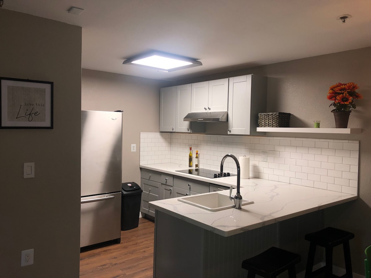 A modern kitchen features sleek gray cabinetry and a large island with seating. Stainless steel appliances, including a refrigerator and stovetop, are present. A minimalist design is accentuated by white tile backsplash and a bright overhead light, creating a functional cooking space.