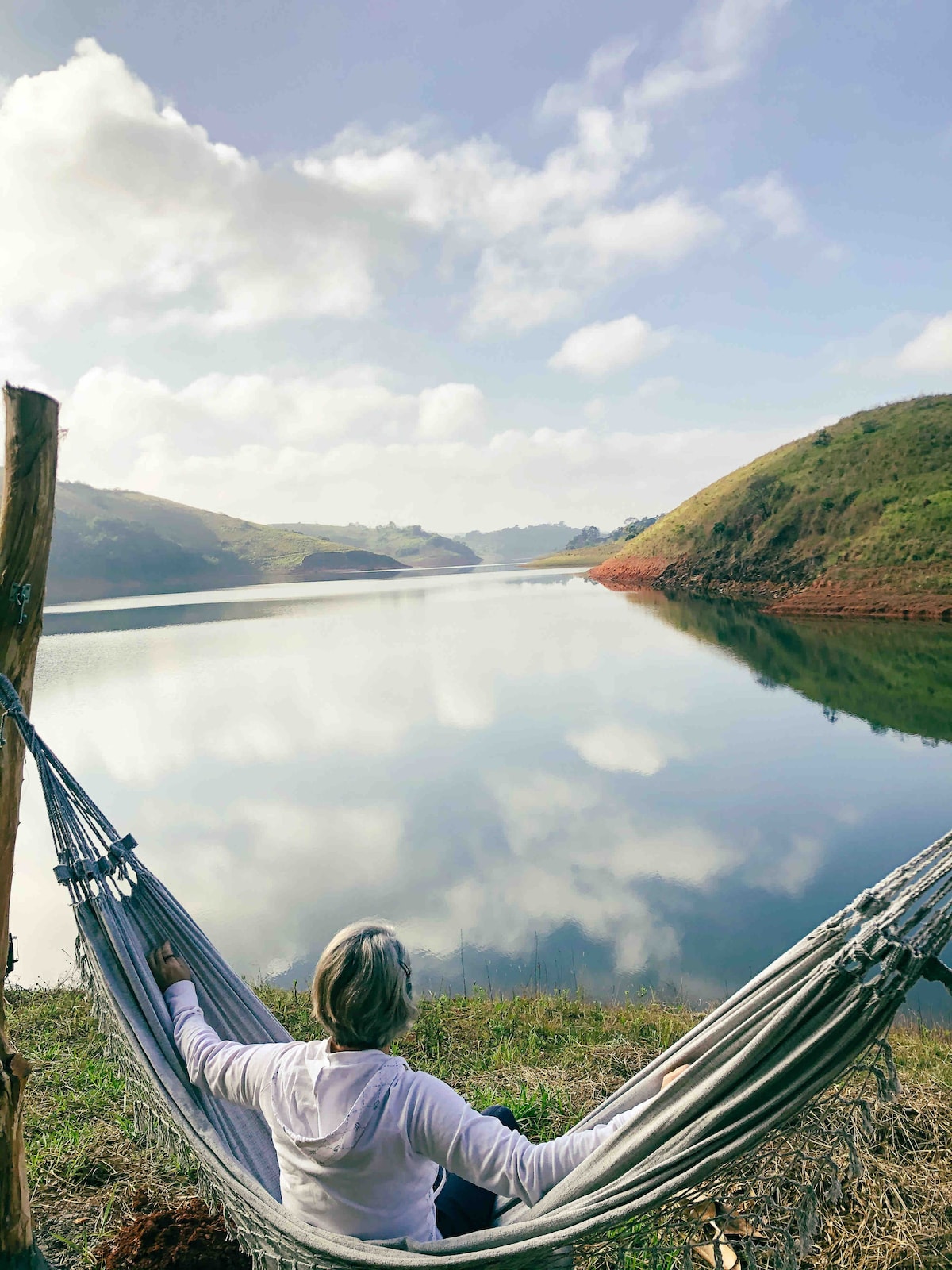 A person relaxes in a hammock by a serene lake, surrounded by green hills. The still water reflects the sky and landscape, creating a calming atmosphere. Soft clouds drift overhead, enhancing the natural setting and inviting a sense of peace.
