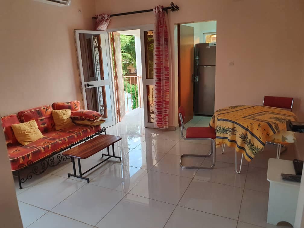 A bright living area is presented, featuring a colorful sofa with accent pillows and a wooden coffee table. A dining table with a patterned tablecloth and red chairs is visible. Large windows allow natural light to fill the space, enhancing the welcoming ambiance.