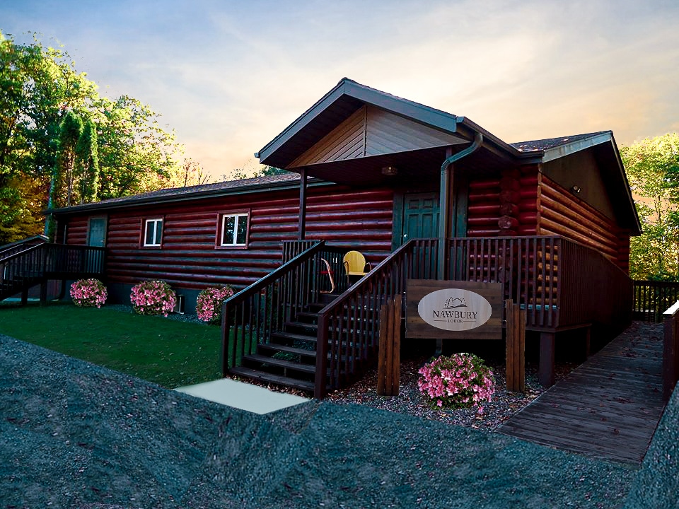 The lodge features a log exterior with a welcoming entrance, complemented by a wooden staircase leading to a spacious deck. Colorful flower beds frame the entrance, while a sign reading 'Nawbury' is prominently displayed, inviting guests to explore the retreat.