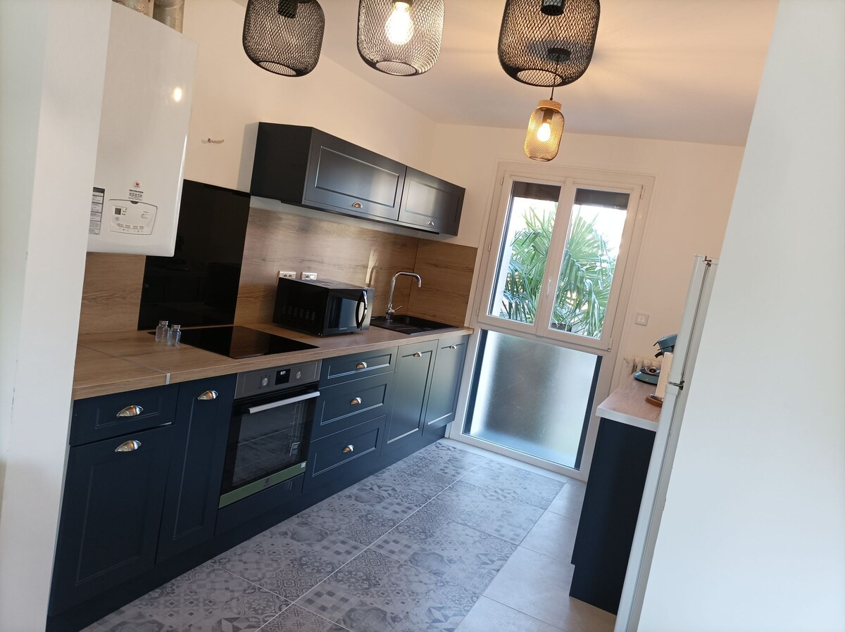 A modern kitchen featuring dark cabinetry and a contrasting wooden countertop. The space is equipped with an oven, stovetop, and sink. Natural light streams in through a large window, illuminating the tiled floor and plants visible outside.