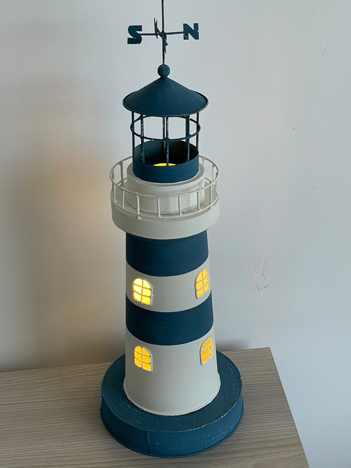 A lighthouse-shaped decor item features a white and blue striped design with small glowing windows. The structure is perched on a light wooden surface, and a compass rose is visible at the top, suggesting nautical themes.