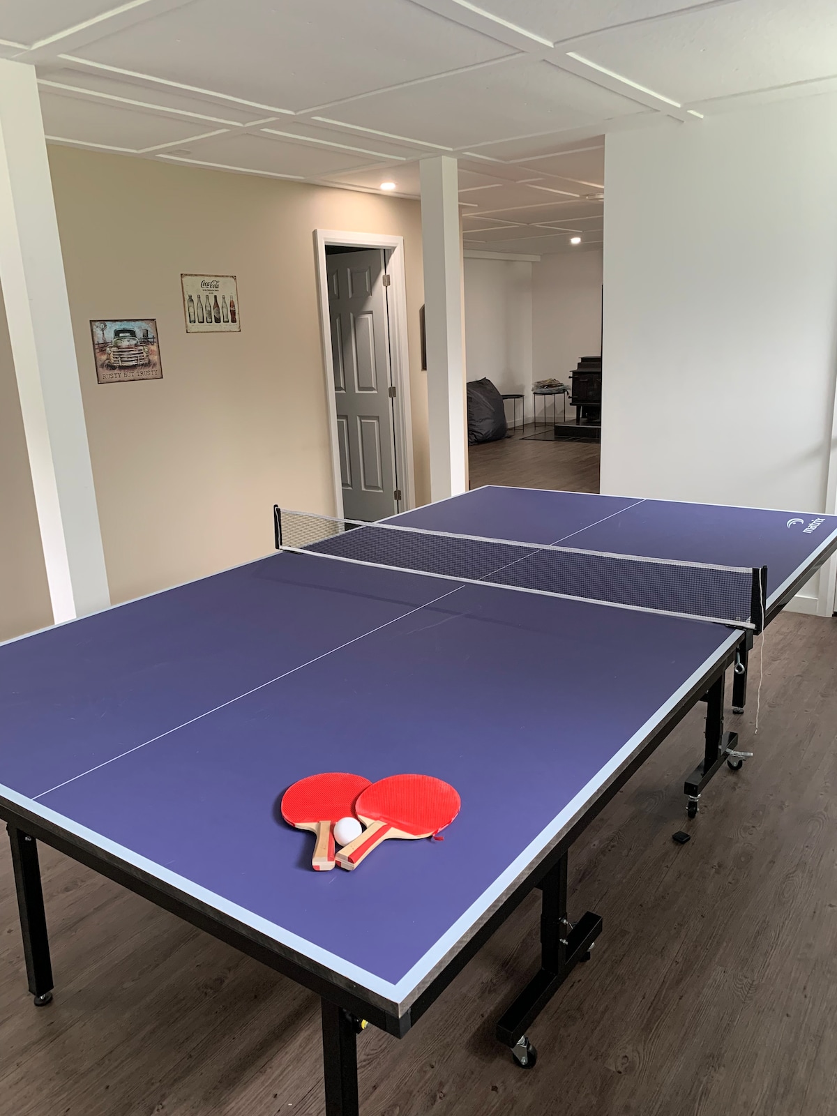 A table tennis setup is displayed in a well-lit recreational space, featuring a blue table with black trim. Two paddles with red handles are placed on the table, inviting playful competition. The background includes a cozy seating area, enhancing the space's leisure appeal.
