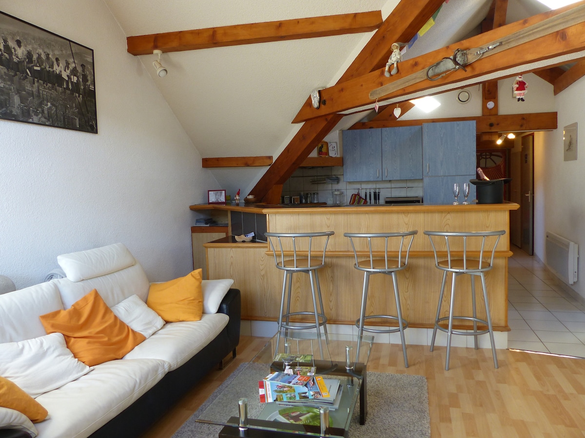 An open-concept living area is depicted, featuring a sofa adorned with orange cushions. A wooden bar with three stools separates the kitchen from the sitting space. Exposed wooden beams add architectural interest to the ceiling, and a coffee table with a decorative display is centered on the floor.
