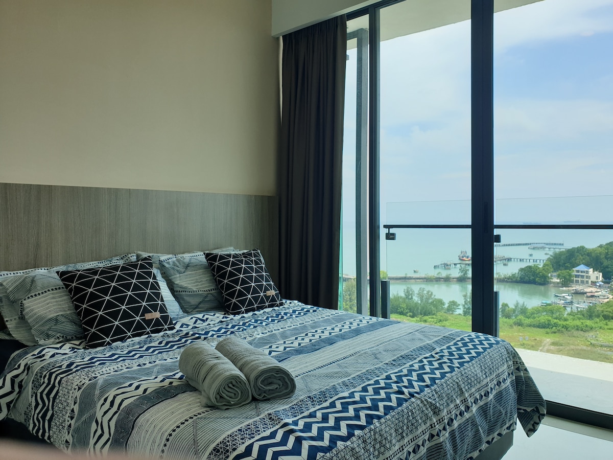 A spacious bedroom features a queen-size bed with a patterned bedspread and two rolled towels at the foot. Large glass doors provide access to a balcony with a view of the water and nearby boats, while dark curtains frame the window.