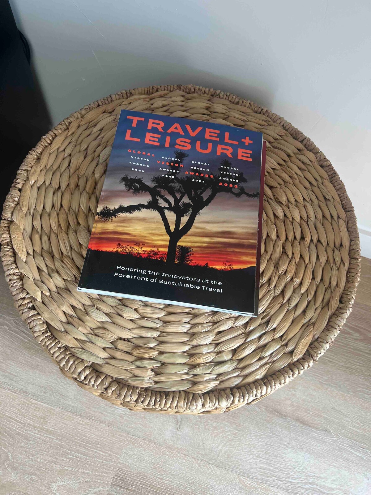 A woven coffee table holds a copy of 'Travel + Leisure.' The cover features a striking image of a tree silhouetted against a colorful sunset, showcasing hues of orange, pink, and deep blue. The table's texture adds a natural element to the space.