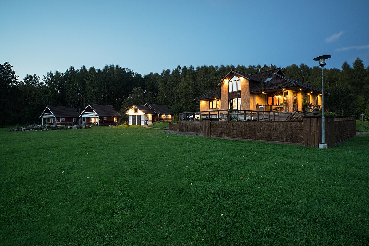A collection of cozy, well-lit cabins and a main building are set against a serene forest backdrop during dusk. Well-maintained green lawn surrounds the structures, and warm lighting from the windows adds to the peaceful ambiance.