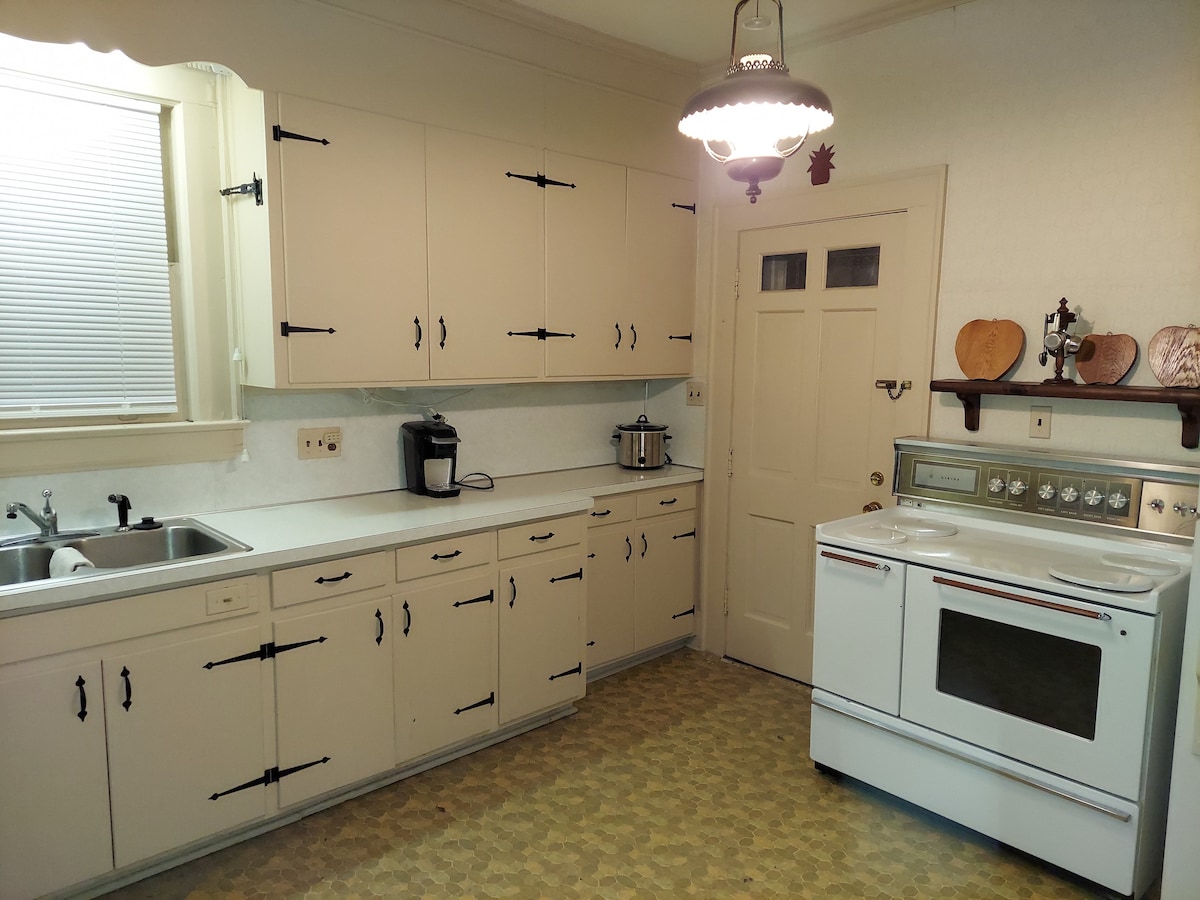 A functional kitchen showcases light-colored cabinetry with contrasting dark hardware. Appliances include a built-in stove and a coffee maker. A window allows natural light to enter, and a door leads to a separate area. The patterned floor adds subtle character to the space.