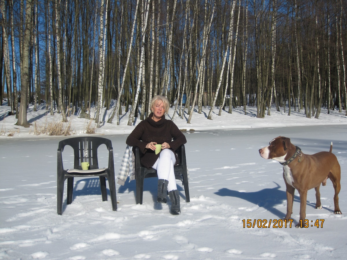 A person sits comfortably in a chair on snow-covered ground. Two chairs are positioned nearby. A friendly dog stands next to the person, with a backdrop of trees and a frozen landscape.