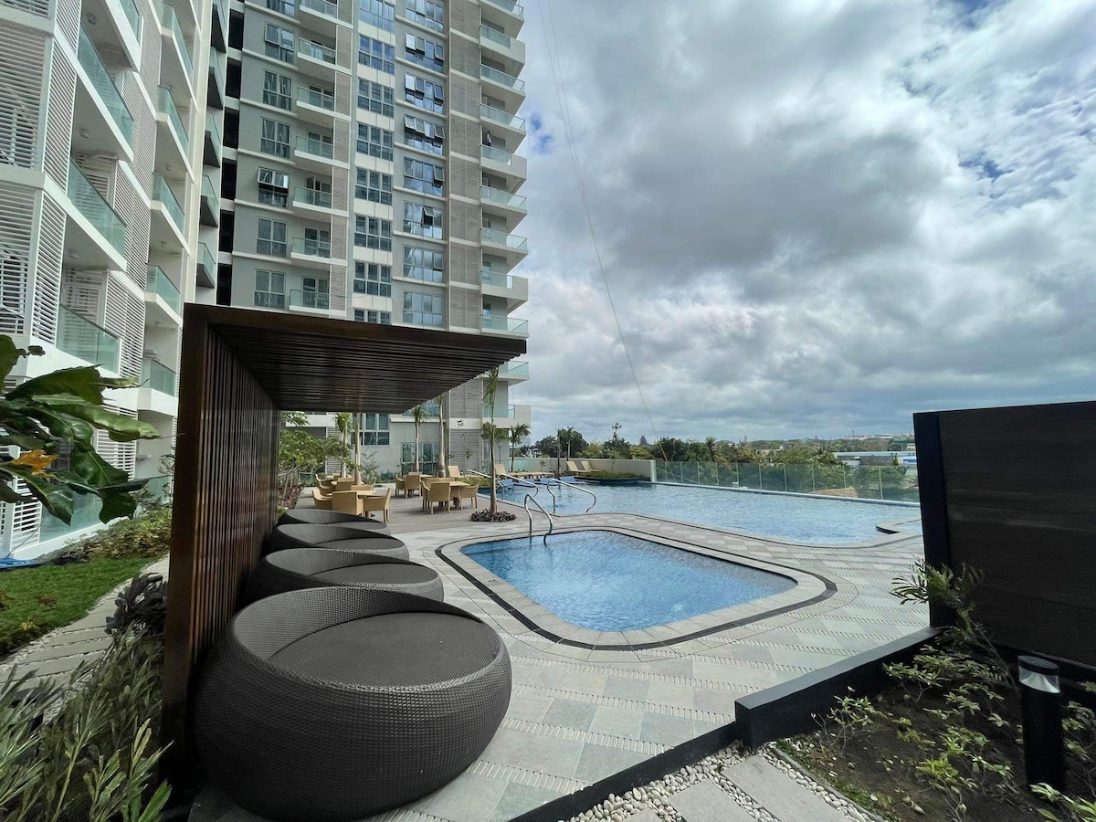 A relaxing outdoor area features three large, rounded chairs and a swimming pool, surrounded by greenery. The pool is partially shaded by a wooden structure, with sun loungers positioned nearby. A modern condominium building stands in the background, visible under a cloudy sky.