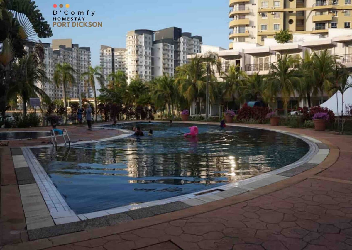 A curved swimming pool is surrounded by palm trees and neatly arranged flower beds. Guests are seen enjoying the water, with areas of the pool reflecting the surrounding architecture. The vibrant foliage adds a touch of greenery to the outdoor space.