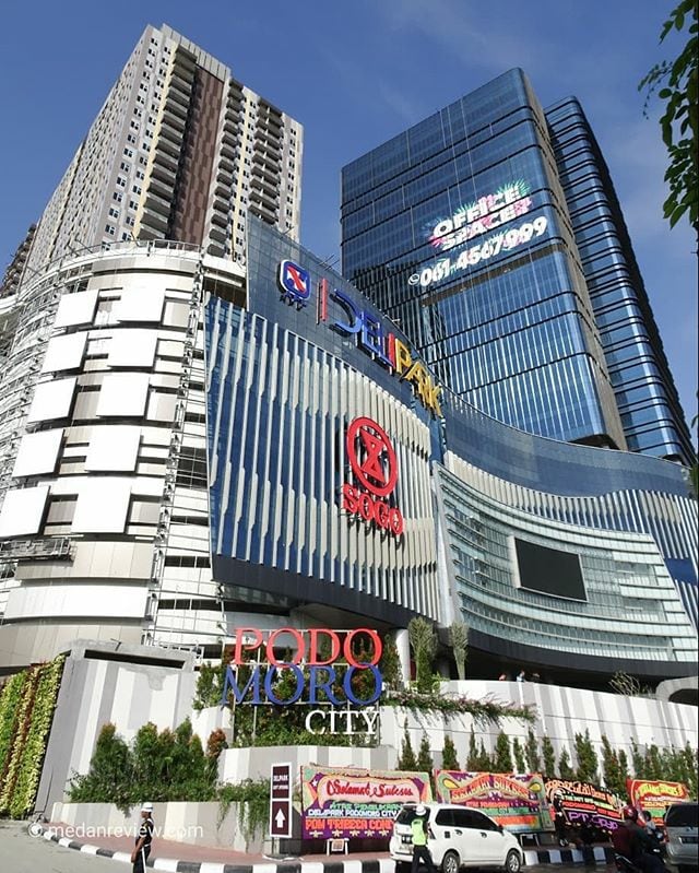 The exterior of Podomoro City Deli Medan is captured, showcasing a modern architectural design with multiple glass facades. Prominent signage indicates Delipark Shopping Mall and Podomoro City, surrounded by landscaped greenery. The structure rises tall against a clear blue sky, reflecting its urban environment.