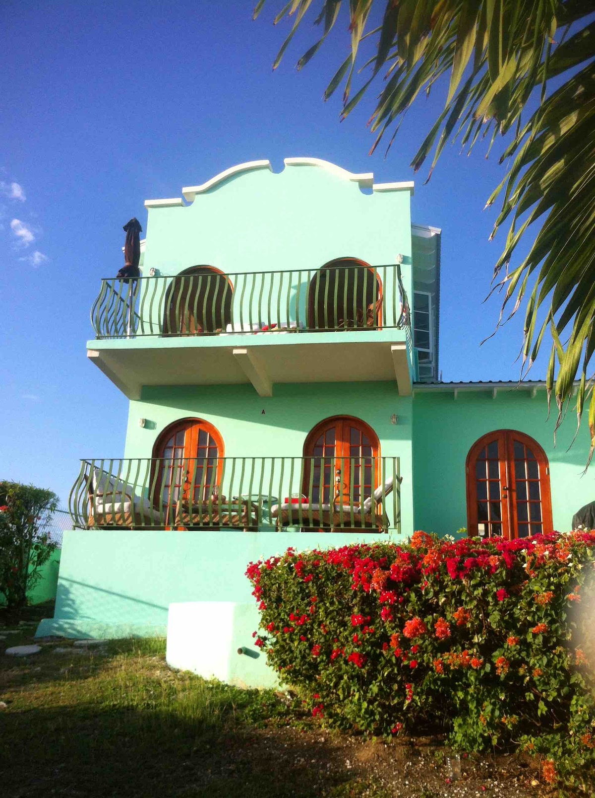 The exterior of the studio apartment features a charming mint green facade. Two balconies with decorative railings are visible, showcasing arched windows that enhance the architectural design. Vibrant bougainvillea plants in front add a touch of color to the landscape.