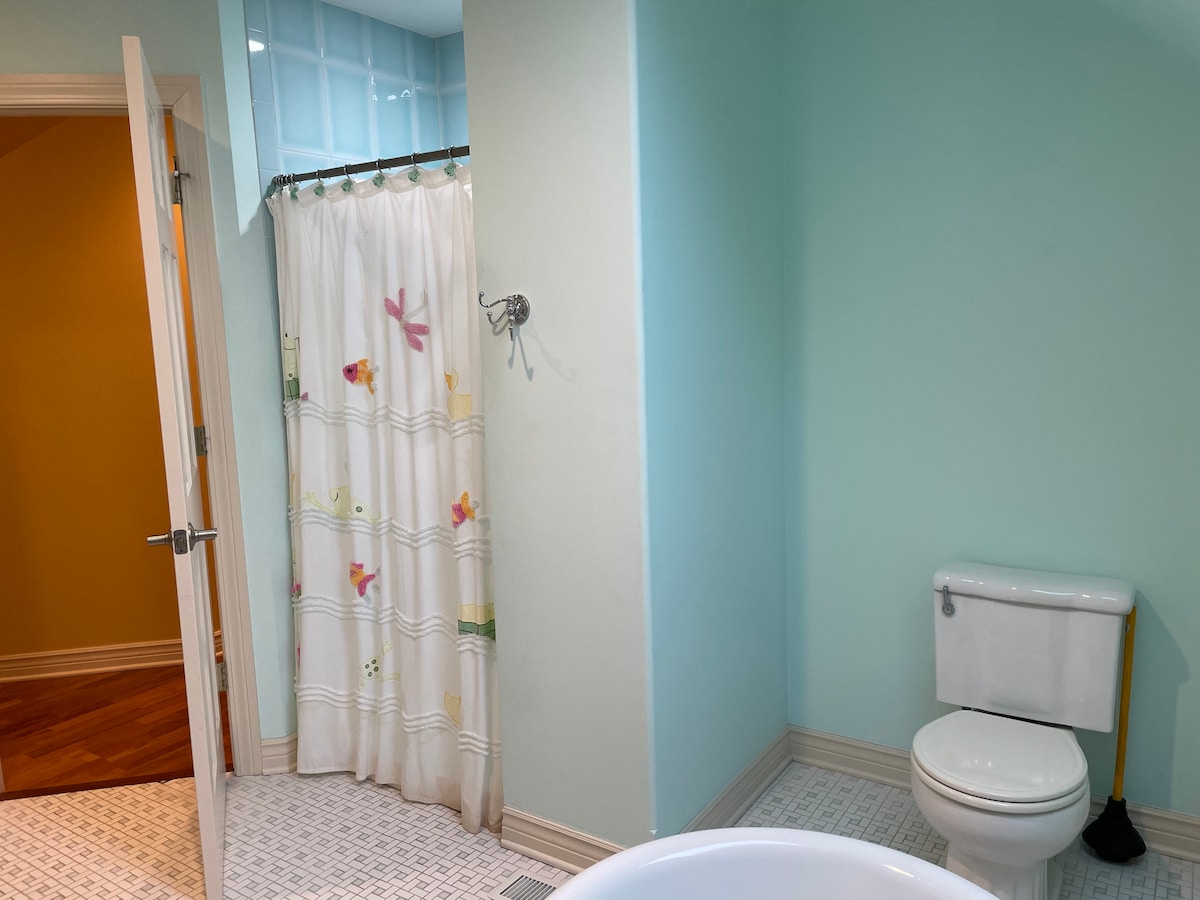 A bathroom is featured with light blue walls and a shower curtain adorned with colorful floral patterns. A spacious bathtub is visible alongside a white toilet. A warm yellow wall can be seen in the adjoining room, adding a pop of color.