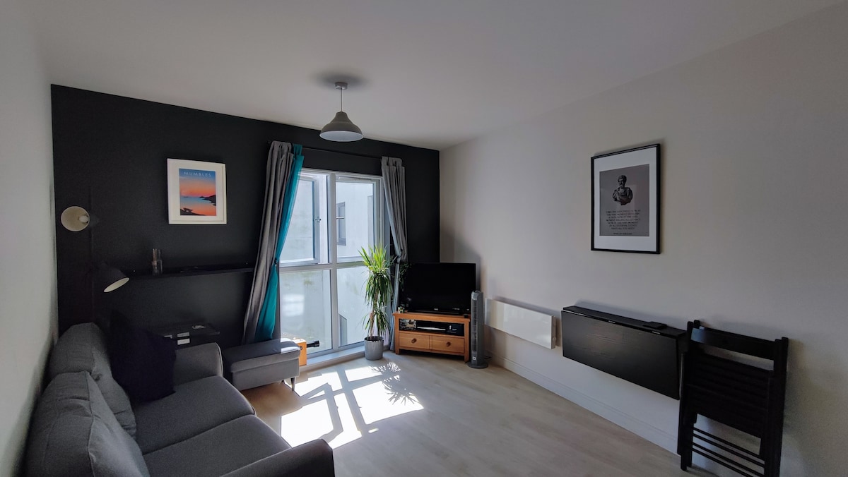 A light-filled living area features a grey sofa and a sleek television. Large windows offer a view outside, framed by curtains. A plant adds a touch of greenery, while framed artwork decorates the walls, enhancing the modern look of the room.