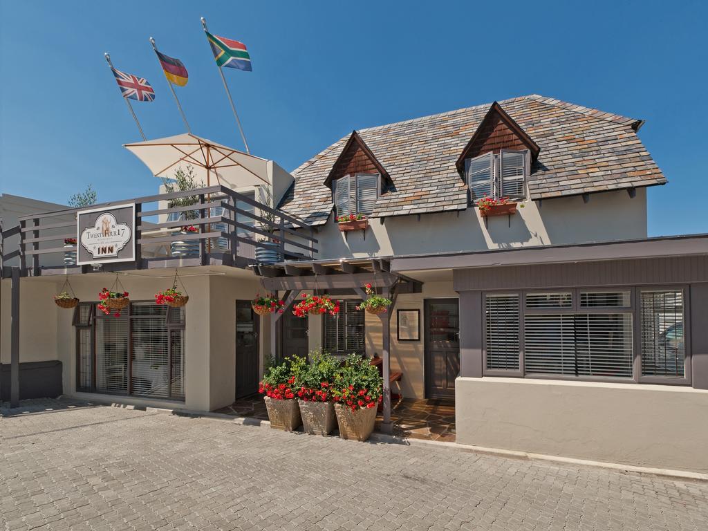 The exterior of the inn features a charming upper balcony adorned with flower pots, along with flags representing the United Kingdom, Germany, and South Africa. The entrance is framed by vibrant red and green flowers, complemented by a tiled pathway and a clear blue sky.