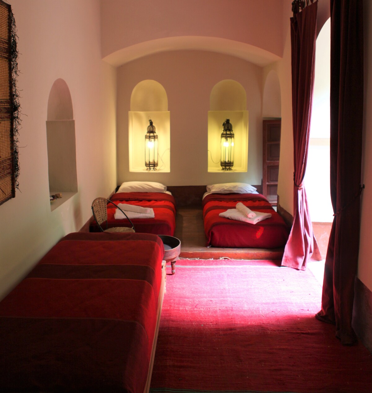 A bright room contains two single beds, each covered with red linens, illuminated by soft lighting from wall sconces. A small table and a woven basket are positioned beside the beds on a warm-toned rug that enhances the inviting space.