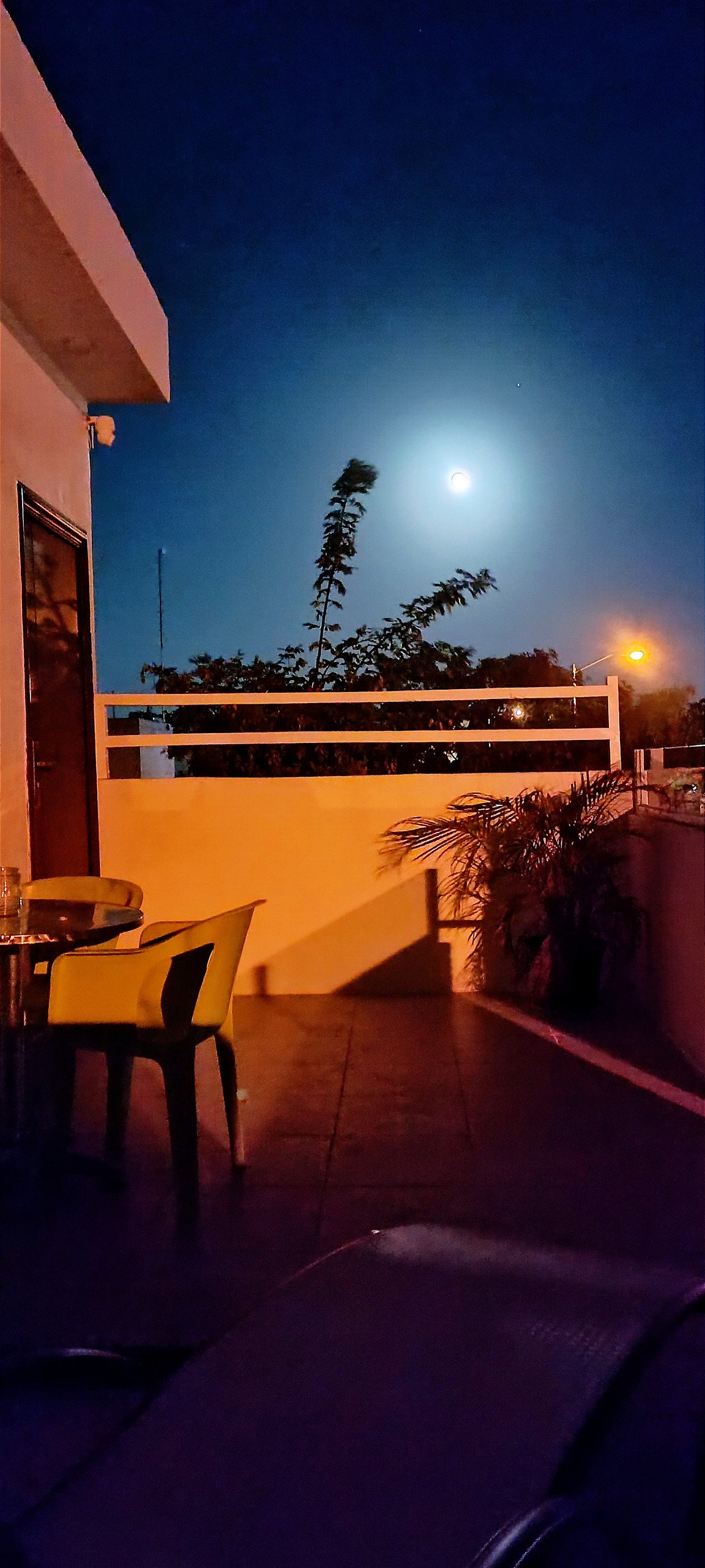 A serene terrace is illuminated by soft moonlight, featuring a small round table and green chairs. Lush foliage is visible, casting gentle shadows, while nearby street lights create a warm ambiance against the night sky.