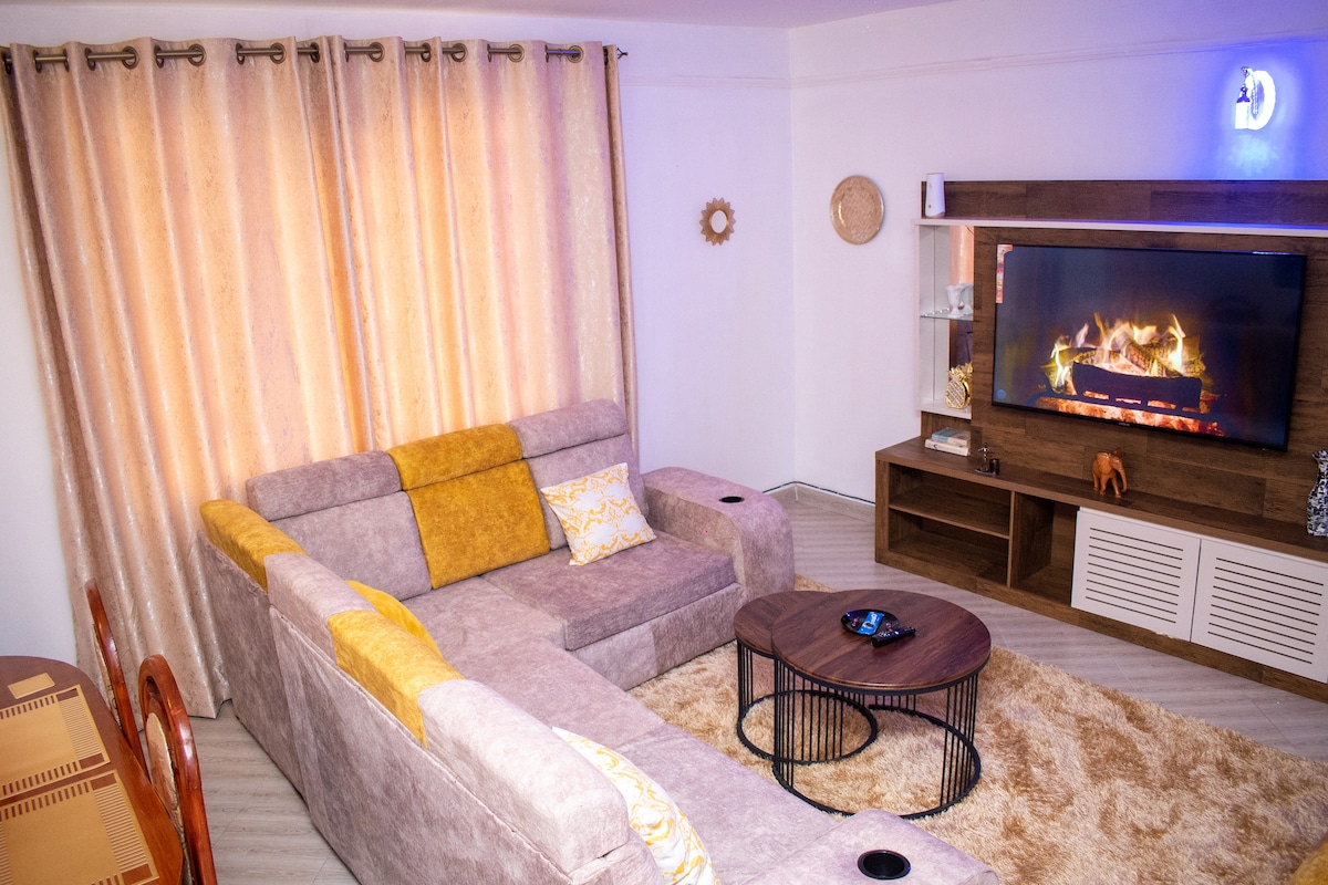A cozy living area features a sectional sofa in neutral tones, complemented by a round coffee table. Soft lighting from floor-to-ceiling curtains highlights a modern entertainment unit with a flat-screen TV above. A plush area rug adds warmth to the space.