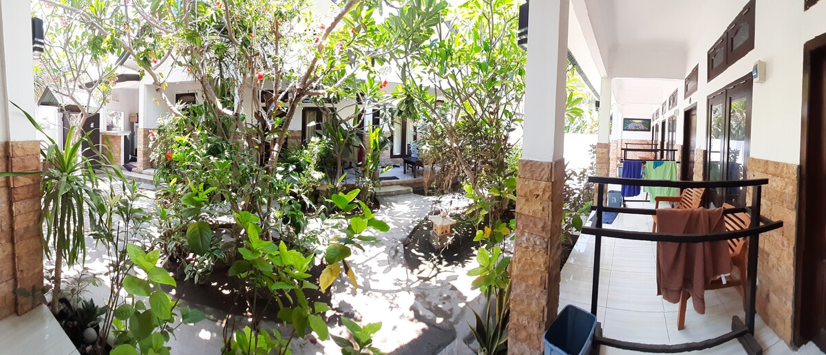 A lush garden area is surrounded by guest rooms, featuring vibrant green foliage and flowering plants. A pathway runs through the garden, leading to individual entrances of the rooms. A drying rack with clothing and a modest seating area are visible on the porch.