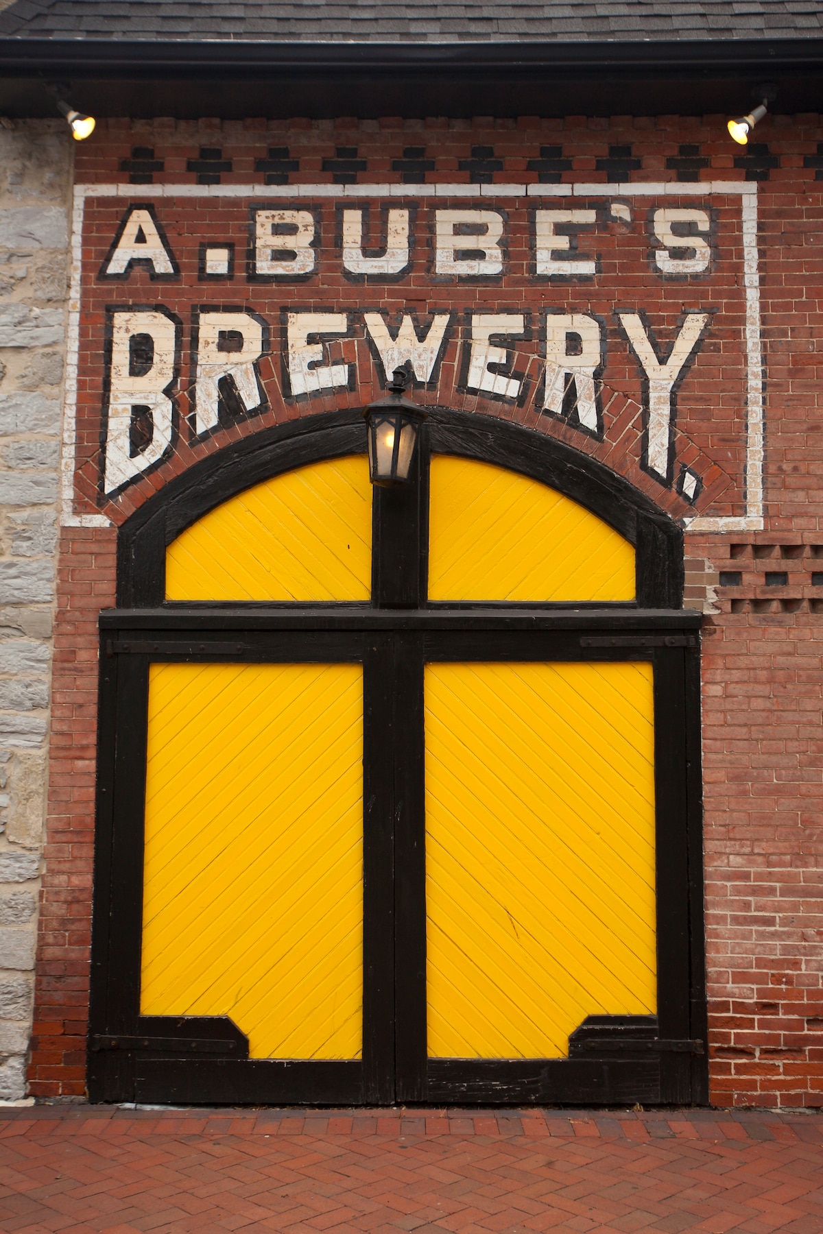 A historic brewery entrance is showcased, featuring large yellow wooden doors set within a striking brick archway. The sign above displays 'A. Bube's Brewery' in bold white lettering against the rustic brick background, complemented by a lantern on either side.