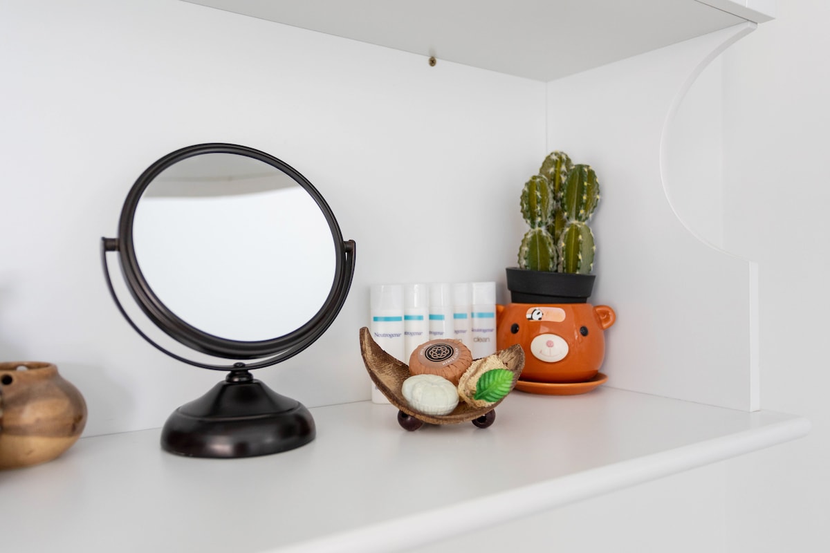 A well-organized shelf displays a round mirror alongside a decorative dish holding soaps. A small cactus plant is housed in a playful ceramic bear container, positioned next to several small bottles. The clean white background emphasizes the colorful accents.