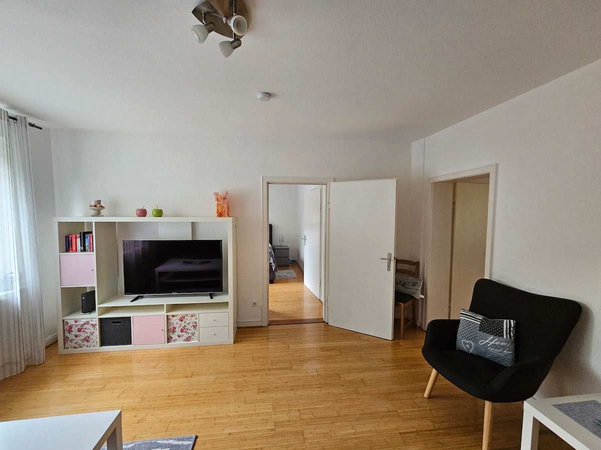 A welcoming living space features a light-colored shelving unit with a flat-screen television atop it. A comfortable chair is positioned nearby, and a doorway leads to a bedroom area in the background. Natural light filters through sheer curtains, enhancing the bright atmosphere.