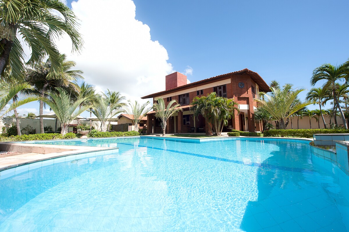 A large swimming pool reflects clear blue skies and is surrounded by palm trees and lush greenery. The two-story house, featuring terracotta accents, stands in the background, providing a relaxing atmosphere for guests.