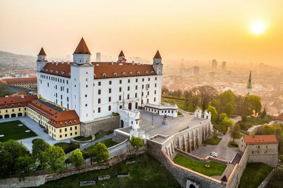 An aerial view showcases the majestic Bratislava Castle with its distinctive tall towers and terracotta roofs. The surrounding landscape features greenery and well-maintained gardens, contrasting with the urban skyline in the background, illuminated by the soft glow of a sunset.