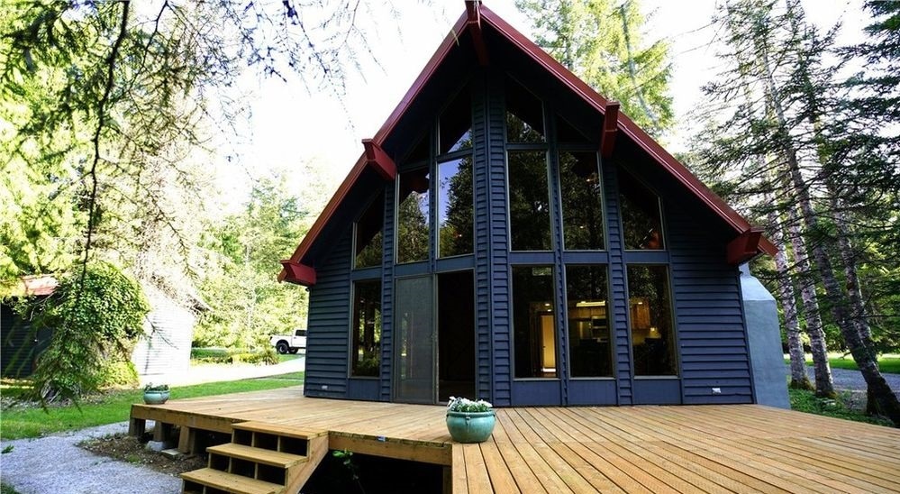 The exterior of the cabin features a steeply pitched roof with large windows allowing natural light to illuminate the interior. A spacious wooden deck is accessible from the front, surrounded by greenery, creating a welcoming entrance to the retreat.