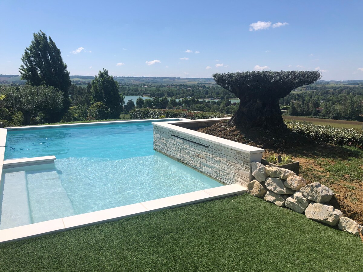 A private, unheated swimming pool is set within a natural landscape, framed by lush greenery and a stone wall. The tranquil blue water mirrors the clear sky, while a large, sculptural tree stands prominently in the background, offering a panoramic view of the surrounding area.