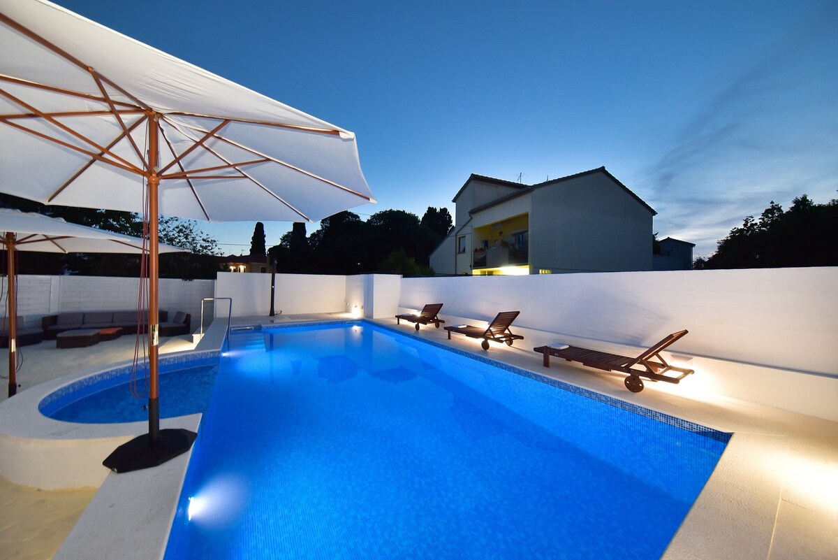 A shared swimming pool is featured, surrounded by sun loungers and large umbrellas. The clear blue water reflects the evening sky as ambient lighting enhances the tranquil atmosphere. The pool area is enclosed by a white wall, offering privacy while enjoying a peaceful outdoor experience.