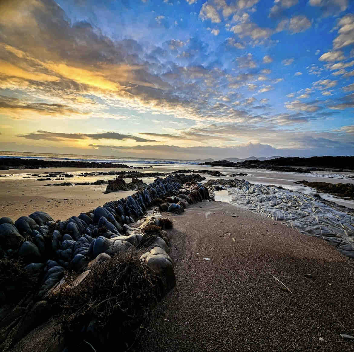 A serene beach scene captures smooth, undulating sand merging with rocky formations at low tide. The sky is painted with soft hues of orange and blue as the sun sets, while gentle waves can be seen lapping against the shore in the distance.