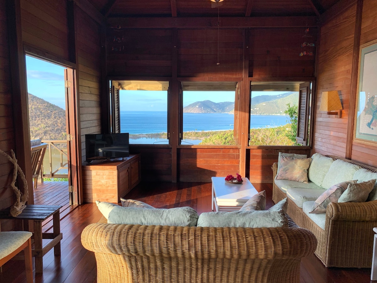 The living area features wooden walls and flooring with panoramic windows offering views of the ocean and nearby hills. A comfortable rattan sofa and matching armchairs surround a coffee table, while a television is positioned against one wall. The space is bright with natural light.