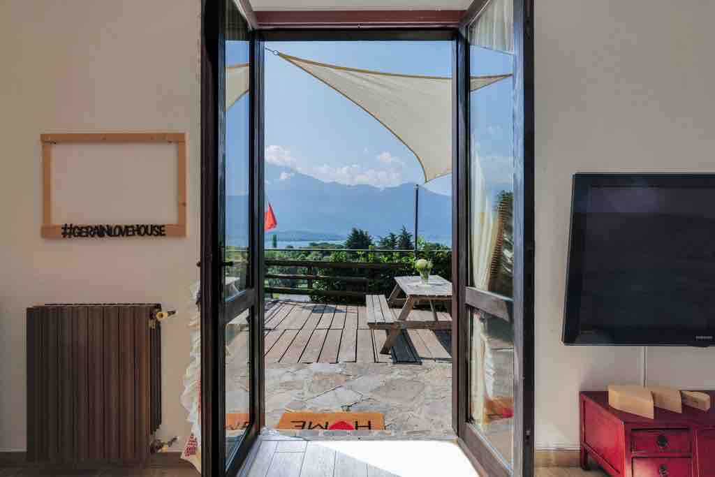 A door opens to a scenic outdoor space, featuring a wooden deck with a picnic table and panoramic views of the mountains and lake. Soft fabric canopies provide shade, enhancing the openness of the area. Natural light floods the interior, creating a bright and airy atmosphere.